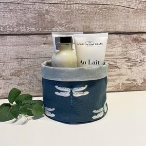 May include: A blue fabric basket with a dragonfly design, containing a bottle and two tubes of Scottish Fine Soaps products. The basket has a pale blue trim. The text 'Au Lait' is visible.
