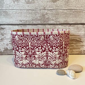 May include: A rectangular fabric storage basket with a burgundy and white pattern featuring birds, rabbits, and foliage. The interior lining has a red and white striped pattern. The basket is on a white surface.