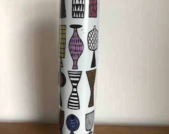Może przedstawiać: Biały ceramiczny wazon o cylindrycznym kształcie z powtarzającym się wzorem stylizowanych waz w kolorze czarnym, brązowym, żółtym, różowym i niebieskim.