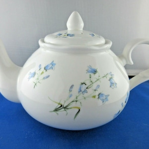 May include: White ceramic teapot with a lid and handle. The teapot is decorated with blue floral designs. The spout and handle are curved. The teapot is sitting on a blue surface.