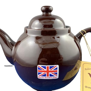 May include: A brown ceramic teapot with a rounded body, spout, and handle. The teapot has a lid with a small knob and a sticker with the Union Jack flag that says "Made in England". A tag reads "Adderley Ceramic" and "Brown Betty Teapot".