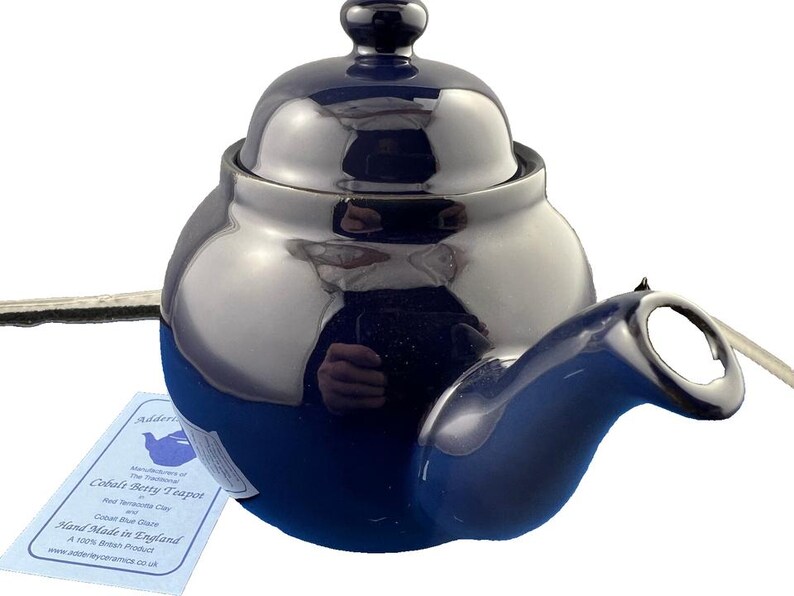 May include: A cobalt blue teapot with a matching lid and handle. The teapot has a glossy finish and a rounded shape. A tag attached to the teapot reads "Cobalt Betty Teapot" and "Hand Made in England".