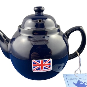 May include: A navy blue ceramic teapot with a glossy finish. The teapot has a rounded body, a curved spout, and a loop handle. A sticker with the Union Jack flag and "MADE IN ENGLAND" is on the front. A tag reads "Adderley Ceramics".