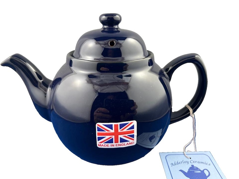 May include: A navy blue ceramic teapot with a glossy finish. The teapot has a rounded body, a curved spout, and a loop handle. A sticker with the Union Jack flag and "MADE IN ENGLAND" is on the front. A tag reads "Adderley Ceramics".