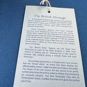 May include: A light blue tag with the text "The British Heritage" and detailed information about the history of Brown Betty teapots. The tag is attached to a string and is set against a blue background.