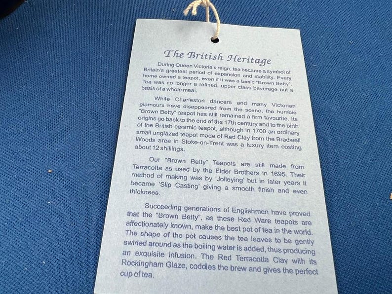 May include: A light blue tag with the text "The British Heritage" and detailed information about the history of Brown Betty teapots. The tag is attached to a string and is set against a blue background.