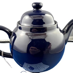 May include: A navy blue ceramic teapot with a glossy finish. The teapot has a rounded body, a curved handle, and a spout. A small lid sits on top, and a tag is attached to the handle. The teapot is designed for brewing tea.