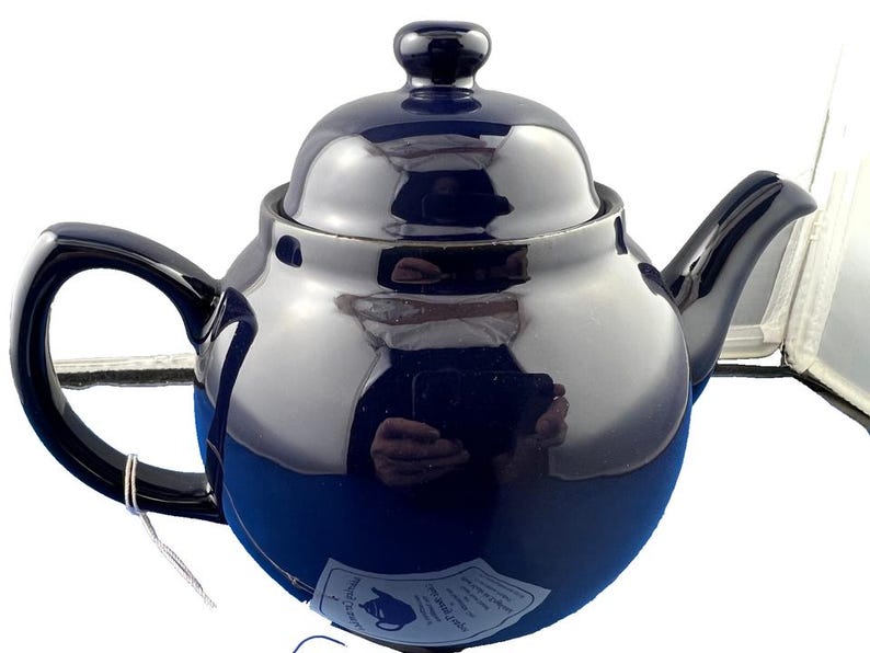 May include: A navy blue ceramic teapot with a glossy finish. The teapot has a rounded body, a curved handle, and a spout. A small lid sits on top, and a tag is attached to the handle. The teapot is designed for brewing tea.