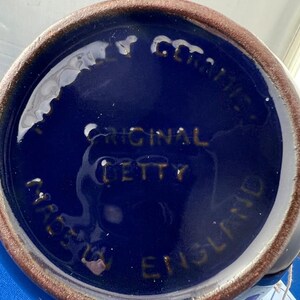 May include: A close-up of the bottom of a blue ceramic mug. The base is dark blue with the words "ORIGINAL BETTY" and "MADE IN ENGLAND" in gold lettering. The rim is brown.