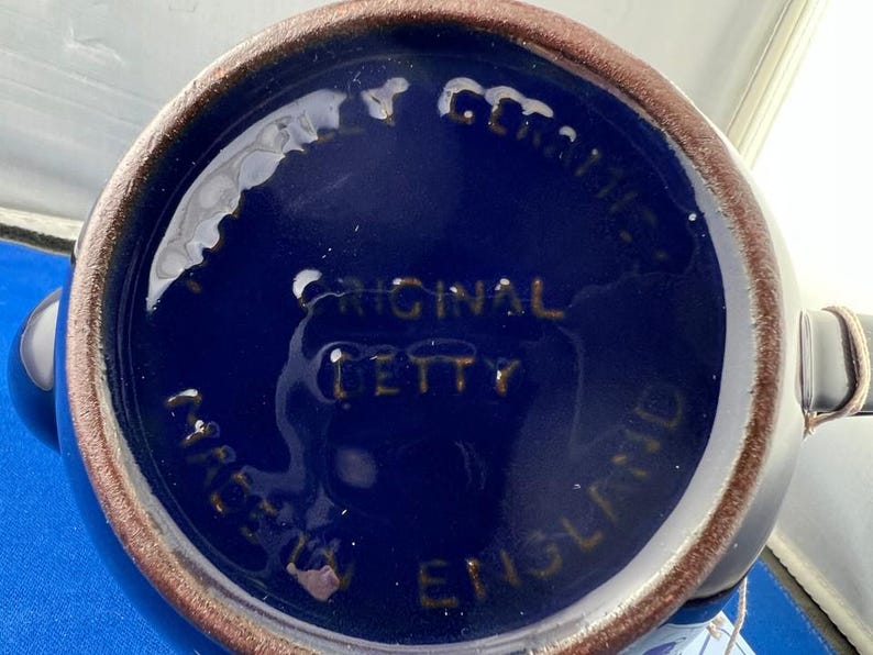 May include: A close-up of the bottom of a blue ceramic mug. The base is dark blue with the words "ORIGINAL BETTY" and "MADE IN ENGLAND" in gold lettering. The rim is brown.