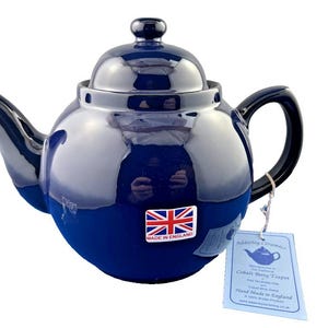 May include: A cobalt blue ceramic teapot with a glossy finish. The teapot has a rounded body, a curved spout, and a black handle. A tag with the text "Cobalt Betty Teapot" and a Union Jack sticker are attached.