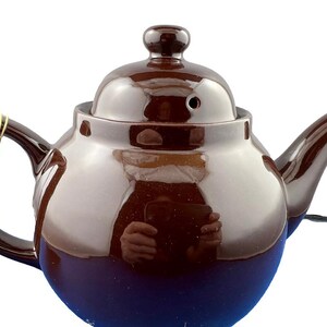 Puede incluir: Una tetera de cer&aacute;mica marr&oacute;n con tapa y asa a juego. La tetera tiene un acabado brillante y una base azul oscuro. Una cuerda dorada est&aacute; sujeta al asa. La tetera est&aacute; dise&ntilde;ada para preparar t&eacute;.