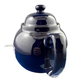 May include: A navy blue ceramic teapot with a glossy finish. The teapot has a rounded body, a curved handle, and a lid with a small knob. The teapot is designed for brewing tea and has a classic, elegant appearance.