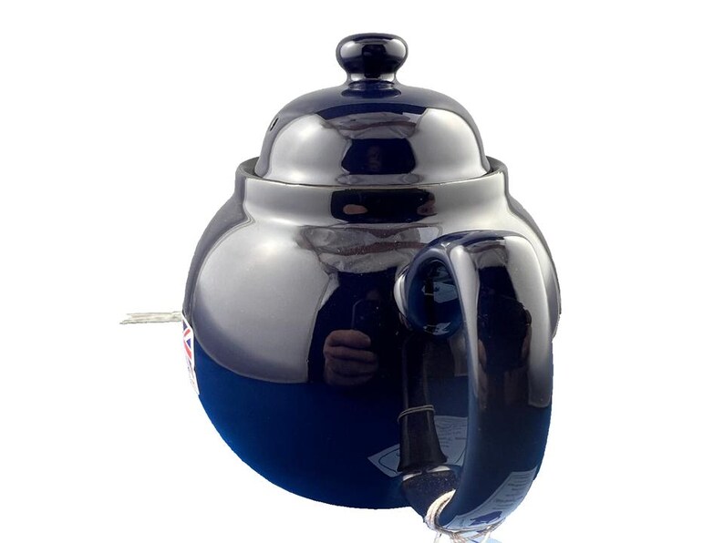 May include: A navy blue ceramic teapot with a glossy finish. The teapot has a rounded body, a curved handle, and a lid with a small knob. The teapot is designed for brewing tea and has a classic, elegant appearance.