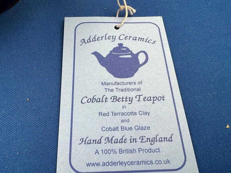 May include: A light blue tag with the text "Adderley Ceramics" and "Cobalt Betty Teapot" in a stylized font. The tag also states "Hand Made in England" and "A 100% British Product". A teapot illustration is also visible.