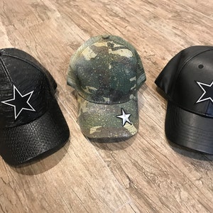 May include: Three baseball caps with star emblems. One is black with a textured pattern, another is camouflage, and the third is solid black. Each cap has a white-outlined star.