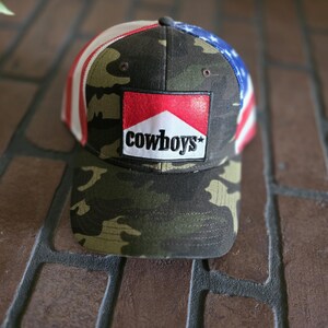 May include: Camouflage baseball cap with a red, white, and blue American flag design on the top. The front of the cap features a patch with the word "cowboys" in white text.