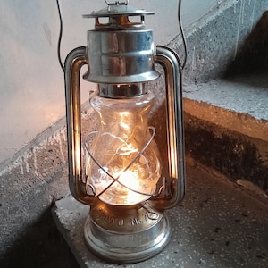 Könnte beinhalten: Eine silberfarbene, antike Öllampe mit einem durchsichtigen Glaszylinder, der von innen beleuchtet wird. Die Laterne, mit einem Griff, ist mit "ROWO No. 10" beschriftet. Sie steht auf einer Betonstufe, was auf eine Verwendung im Freien hindeutet. Das warme Licht kontrastiert mit den kühlen Tönen.