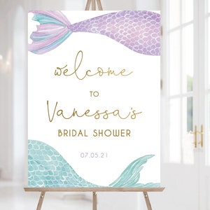 May include: Welcome to Vanessa's Bridal Shower sign with a purple and blue mermaid tail on each side. The sign is white with gold lettering and the date 07.05.21.