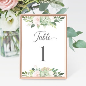 Puede incluir: Una tarjeta de número de mesa blanca con marco dorado y diseño floral verde y rosa. La tarjeta dice "Mesa 1" en fuente gris.