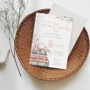 K&ouml;nnte beinhalten: Eine wei&szlig;e Einladung mit rosa und gr&uuml;nen floralen Akzenten. Der Text lautet "One for the Books" und enth&auml;lt Details f&uuml;r eine erste Geburtstagsfeier. Die Einladung zeigt einen Stapel B&uuml;cher, die mit einem rosa Band zusammengebunden sind.