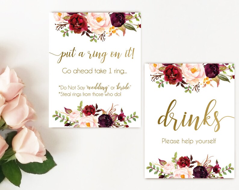 K&ouml;nnte beinhalten: Zwei bedruckbare Schilder mit einem Blumendesign in den Farben Burgunderrot, Rosa und Gr&uuml;n. Das obere Schild sagt "put a ring on it! Go ahead take 1 ring... "Do Not Say 'wedding' or 'bride'." "Steal rings from those who do!" Das untere Schild sagt "drinks Please help yourself"