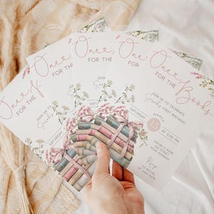 K&ouml;nnte beinhalten: Rosa und wei&szlig;e Einladung zu einem ersten Geburtstag mit dem Text "One for the Books" und einem Stapel B&uuml;cher mit einem rosa Band. Die Einladung enth&auml;lt Datum, Uhrzeit und Ort der Party.
