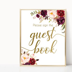 K&ouml;nnte beinhalten: Goldenes Schild mit burgunderrotem und rosafarbenem Blumendesign. Das Schild lautet "Bitte unterschreiben Sie das G&auml;stebuch".
