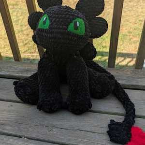 Può includere: Un peluche a forma di drago nero fatto a mano all'uncinetto, con occhi verde brillante e una punta della coda rossa. Il drago ha grandi orecchie arrotondate ed è seduto su una superficie di legno. Il peluche è realizzato con filato morbido e strutturato.