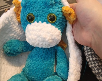 Puede incluir: Un juguete de peluche de dragón de color azul turquesa tejido a crochet con detalles en naranja y grandes ojos negros. El dragón está sentado dentro de un nido tejido a crochet blanco.