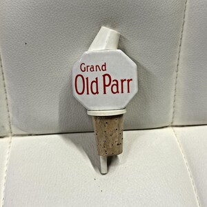 May include: A white ceramic bottle stopper with a cork base. The stopper has a white spout and a white octagonal face with the words "Grand Old Parr" in red lettering. The cork is a light brown color.