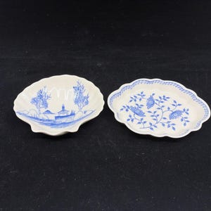 May include: Two small, white porcelain dishes with blue designs. One is shell-shaped with a landscape scene, the other is oval with a floral pattern and a blue scalloped edge. Both are on a dark surface.