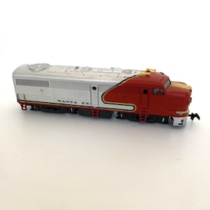May include: A model train locomotive in silver, red, and yellow. The words "SANTA FE" are printed on the side of the train. The train has a streamlined design and is set against a white background.