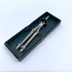 May include: A silver-toned drafting compass with a black adjustment wheel and a sharp point, resting inside a dark green rectangular box. The compass is designed for precise measurements and drawing circles. The box has a worn appearance.