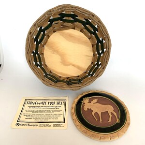 May include: A woven basket with a wooden base and green accents, alongside a stoneware food tile with a moose design. A paper label reads "STONEWARE FOOD TILE!" and provides care instructions. The tile is approximately 6 inches in diameter.