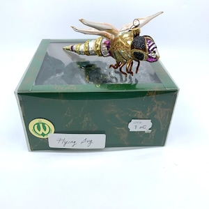 May include: A decorative glass flying bug ornament with gold, purple, and white accents, displayed in a green box. The ornament has delicate wings and intricate detailing. The box has the text "Flying Bug".