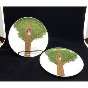 May include: Two white ceramic plates featuring a detailed illustration of a brown tree trunk with green leaves and a small owl perched inside a hollow in the center. One plate is displayed upright on a black stand, while the other lies flat.