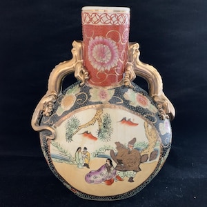 May include: An antique ceramic vase with a red, gold, and black color scheme. The vase features a cylindrical neck with a floral design and two gold handles. The body of the vase is round with a painted scene of figures in a landscape.