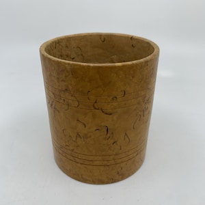 May include: A cylindrical wooden cup with a light brown finish. The cup has horizontal etched lines and decorative markings. The cup is empty and sits on a white surface.