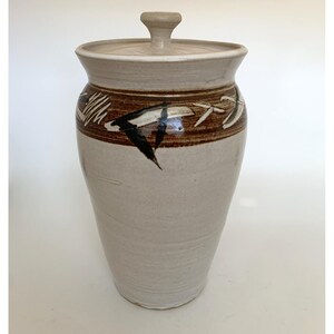 May include: A tall, cylindrical ceramic canister with a matching lid and a knob handle. The vessel features a light beige base glaze with a wide, dark brown horizontal band near the top, decorated with abstract, hand-painted black and white brushstrokes.