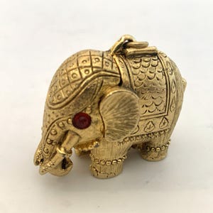 May include: A gold-toned metal elephant charm featuring a textured surface with a grid pattern on the head and scale-like details on the back. The elephant has a single red circular stone set as an eye and a small loop at the top for attachment.