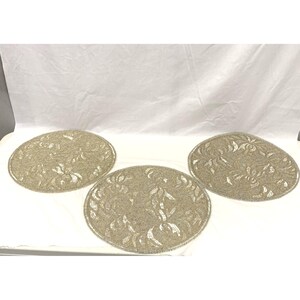 May include: Three round placemats featuring a dense, shimmering silver beaded surface. Each placemat displays a repeating floral pattern created with reflective, metallic-toned beads that catch the light, arranged in a circular, flat design.