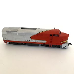 May include: A red, silver, and white Santa Fe model train. The train has the words "SANTA FE" printed on the side. The train is a detailed replica of a diesel locomotive.