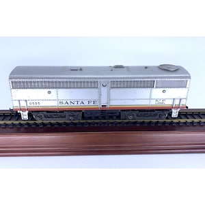 May include: A silver and gray model train car with the words "SANTA FE" and the number "0535" printed on the side. The model train car is on a wooden base with train tracks.