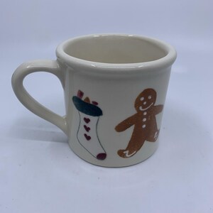 May include: A cream-colored ceramic mug featuring a hand-painted design of a white Christmas stocking with small red hearts and a dark green cuff, alongside a brown gingerbread person figure with white icing details on the front surface.