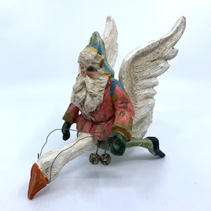May include: A whimsical figurine of Santa Claus riding a goose with large white wings. Santa wears a red coat, blue hat, and green gloves. The goose has an orange beak and two small bells hanging from its neck. A unique Christmas decoration.