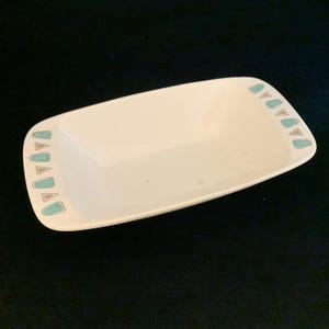 May include: A rectangular, off-white ceramic dish with rounded corners. The dish is decorated with a repeating pattern of teal and brown triangles along the edges. The dish is empty and sits on a dark surface.