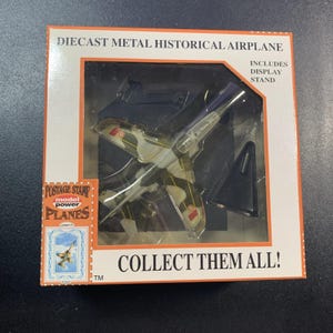May include: Diecast metal historical airplane model in a box. The model has a camouflage design with red and yellow accents. The box reads "Collect Them All!" and includes a display stand.