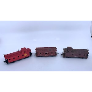 May include: Three model train cars in red and brown. The red car has a Santa Fe logo. The brown cars have details like windows and doors. These miniature train cars are likely for a model railroad or train set.
