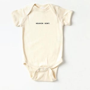 Op de afbeelding: Een cr&egrave;mekleurige baby romper met korte mouwen en de tekst "HEAVEN SENT." in het zwart gedrukt.